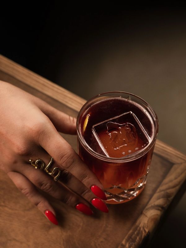 Hand with red nails and snake ring beside a cocktail with a large ice cube.