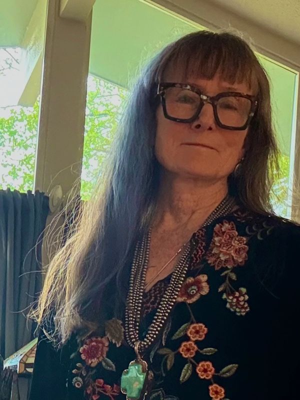 Older woman with long hair and large glasses wearing a floral embroidered top indoors.