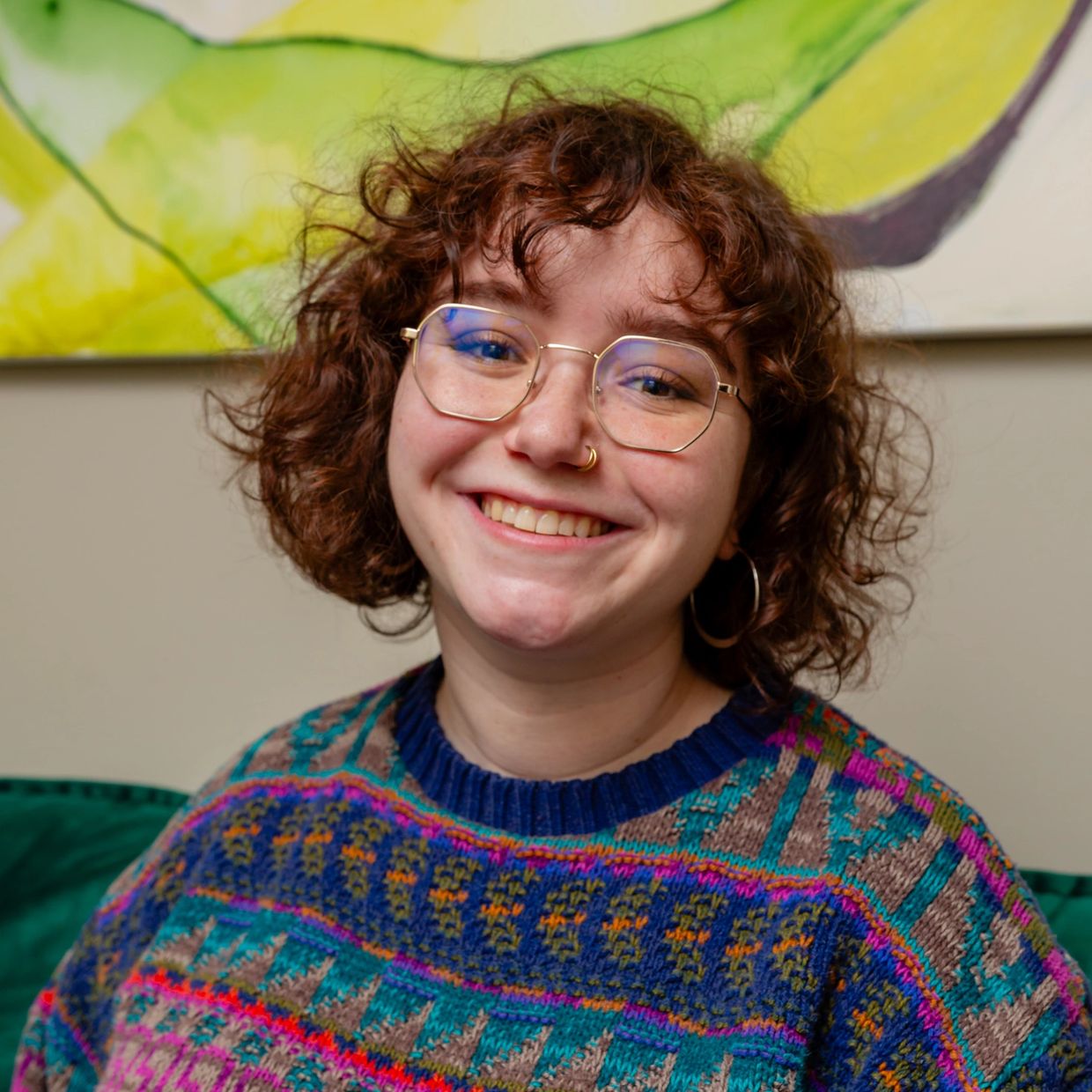 Smiling person with curly hair and glasses wearing a colorful sweater.