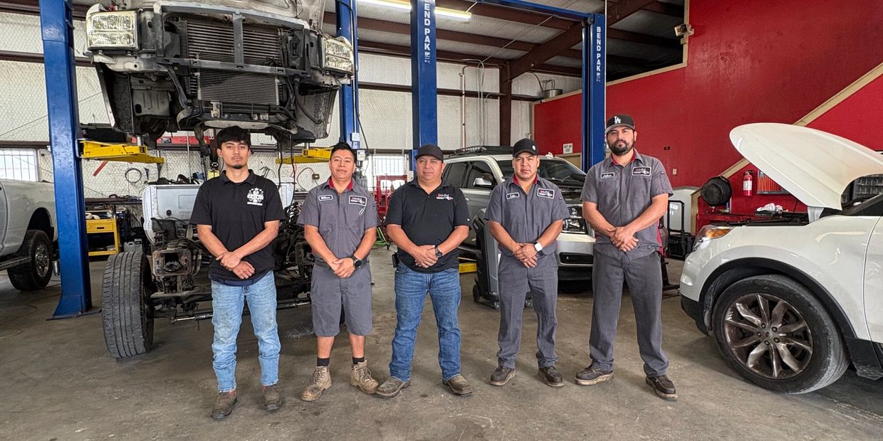 Five auto mechanics standing in a garage with vehicles and equipment.
