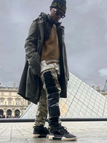 Man in streetwear standing near Louvre Pyramid in Paris on a cloudy day.