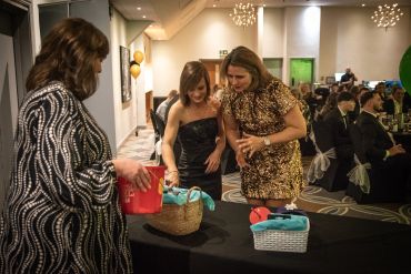 Three women at a formal event examining gift baskets on a table.