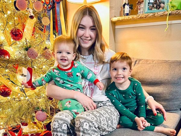 Woman holding two young boys in lap while wearing Christmas pajamas in front of a Christmas tree.