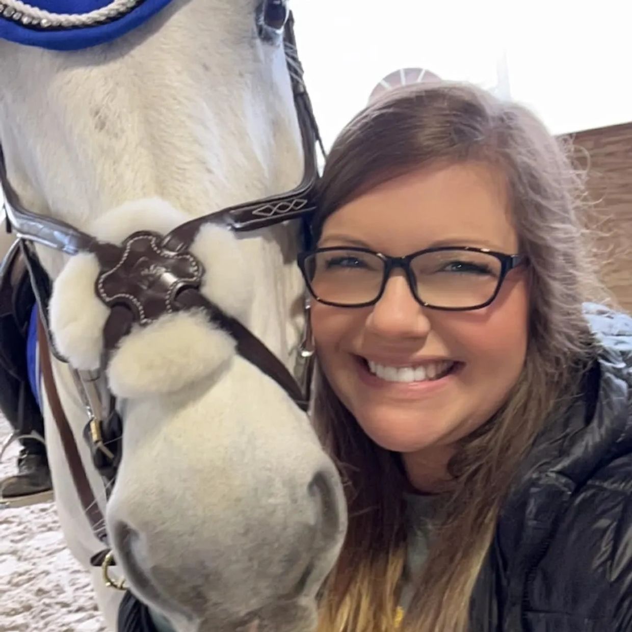 Woman in glasses hugging head of gray horse wearing a blue ear bonnet.