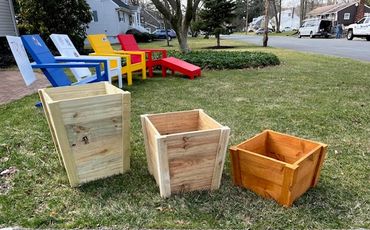 Three wooden planters of different sizes on grass with colorful chairs in background.