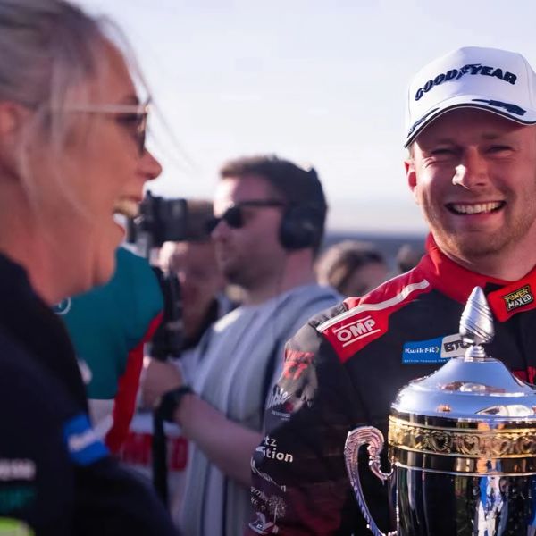 A race car driver smiling with a trophy in hand and people in the background.