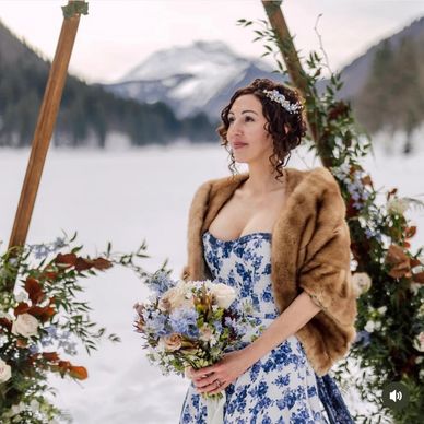 Bride in a floral dress and fur shawl holding a bouquet in a snowy mountain setting.