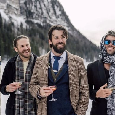 Men in winter attire holding drinks, smiling outdoors in snowy mountains.