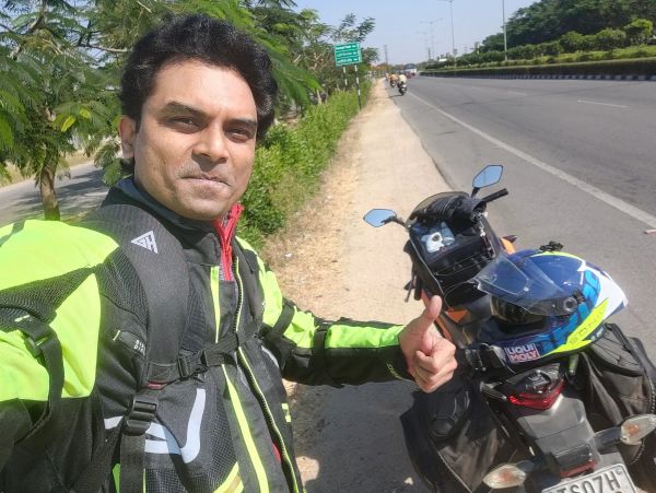 Man in green jacket giving thumbs up beside his motorcycle on a sunny road.