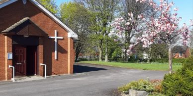 Small brick church with white cross and blooming tree.