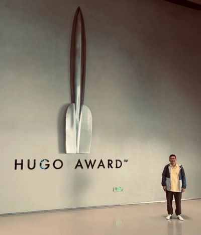 Person standing beside a large Hugo Award sculpture on a wall.