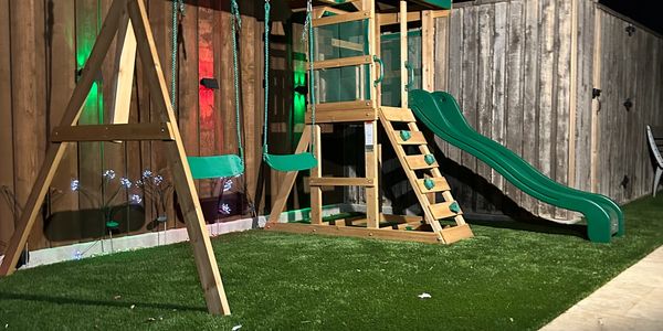 Wooden playground set with swings and slide on artificial grass at night.