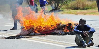 Man sitting beside a flaming mattress on the street.