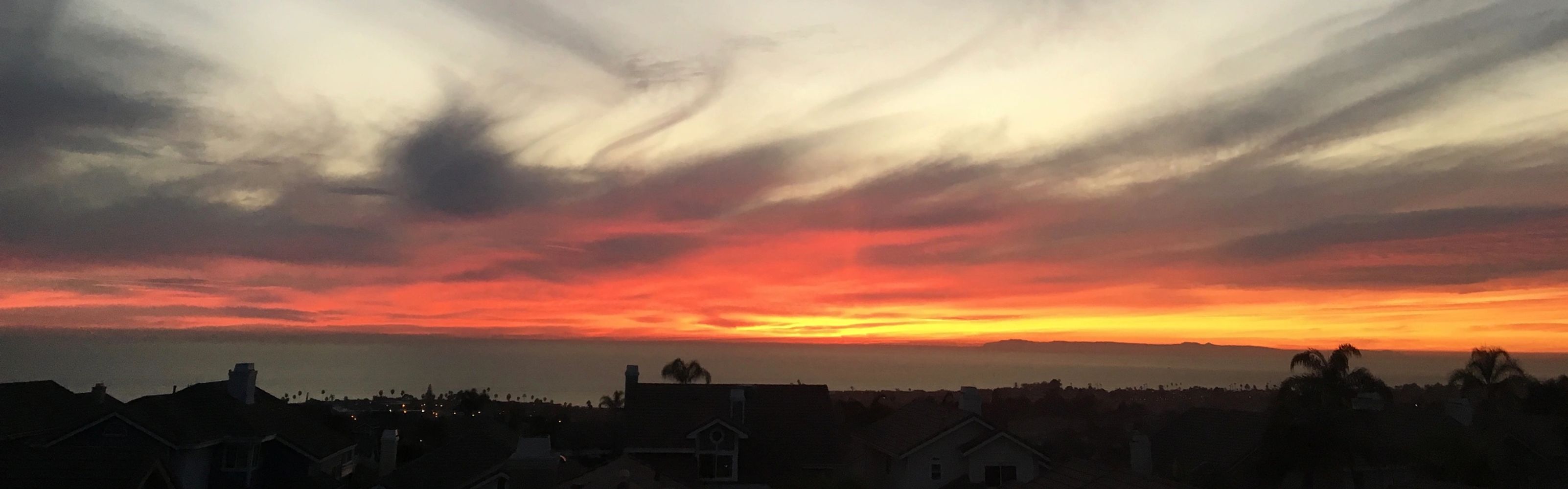 A vibrant sunset with red and orange hues over silhouetted rooftops and palm trees.