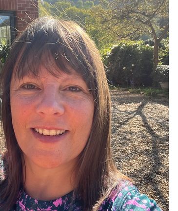 Woman smiling outdoors with sunlight and greenery in background.