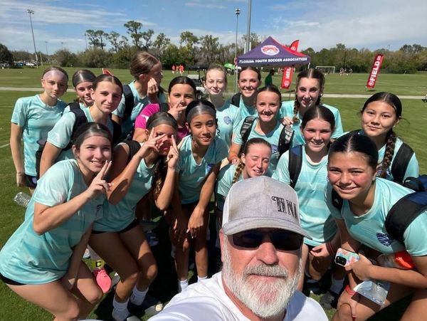 A girls' soccer team and coach posing for a group selfie on a sunny field.