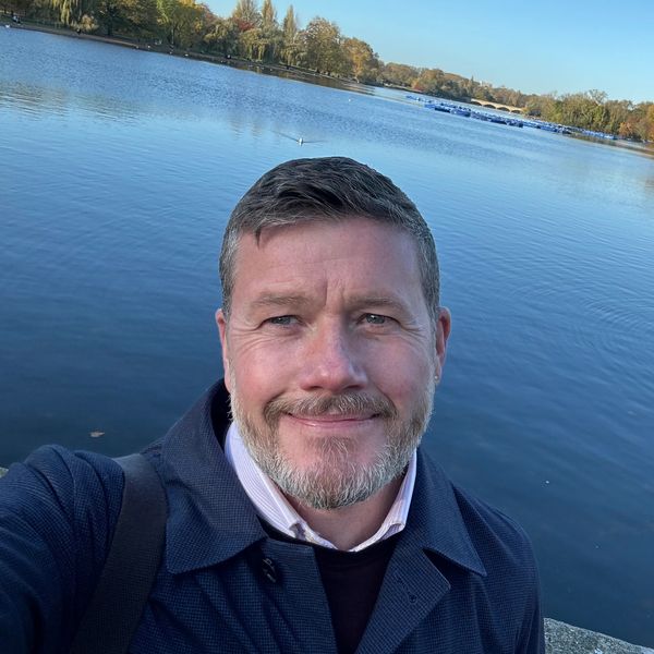 Man smiling for a selfie by a calm lake on a clear day.