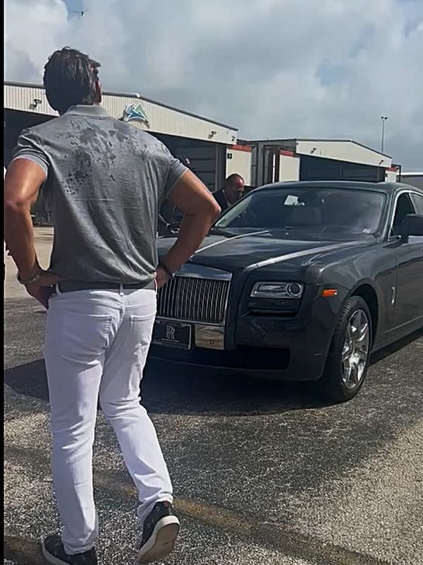 Man in white pants and gray shirt stands near a luxury car.