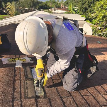 Roofer securing metal bracket on roof with drill, wearing safety gear.