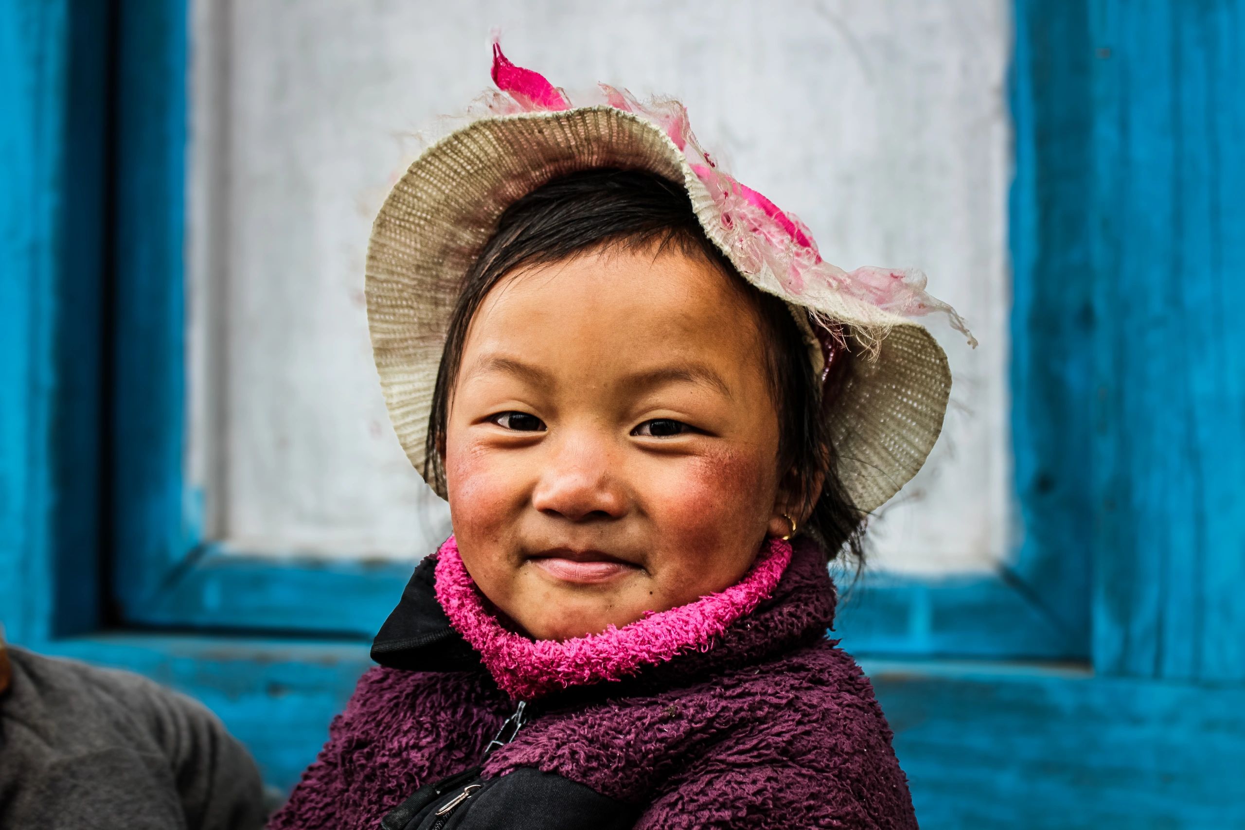 Smiling child wearing a tattered hat and warm clothing in front of a blue window frame.