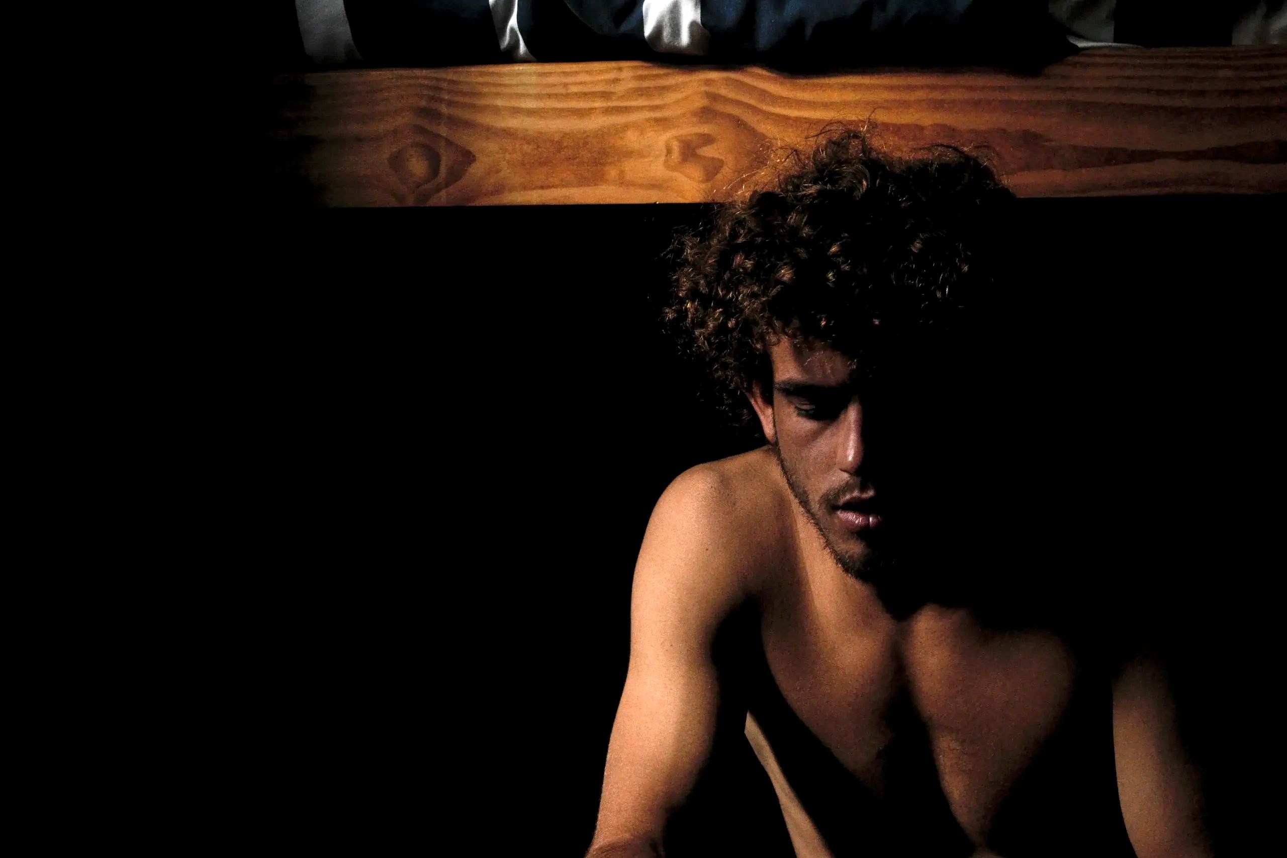 A shirtless man with curly hair in low light, deep in thought.