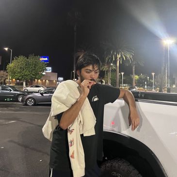 A young man smoking while leaning on a white vehicle at night.
