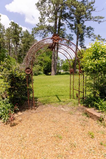 Rustic metal garden archway surrounded by greenery on a sunny day.