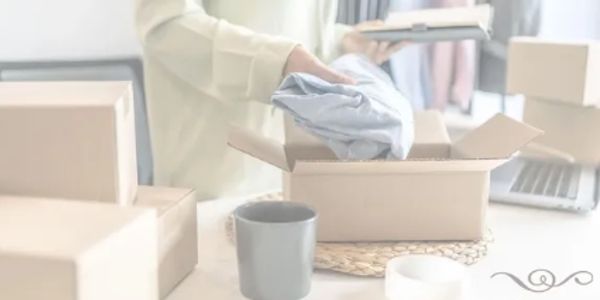 Person packing clothing into a cardboard box surrounded by other boxes and a laptop.
