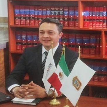 Man in suit sitting at desk with Mexican and institutional flags.