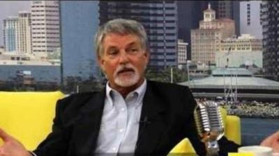 Man speaking on a TV talk show set with city backdrop.