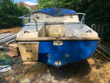 An old, weathered blue and white boat on land surrounded by trees and materials.