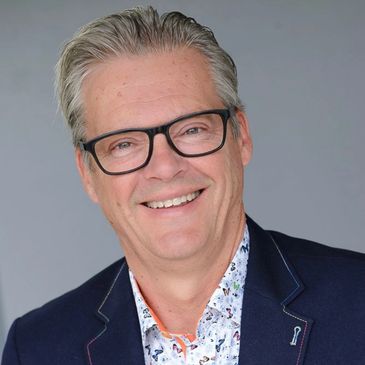 Smiling man with glasses in a patterned shirt and navy blazer.