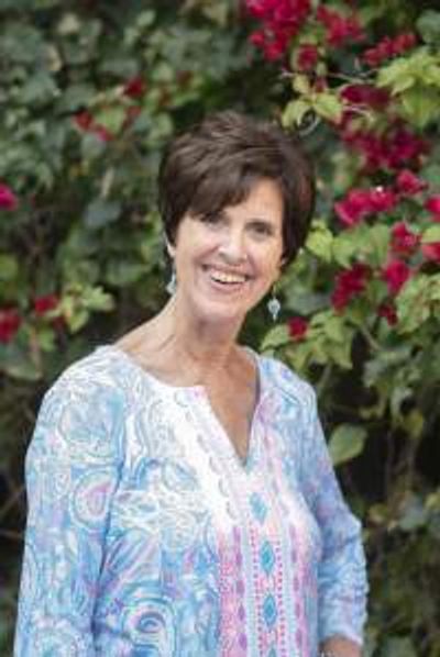 Smiling woman in a colorful blouse stands in front of lush, flowering bushes.
