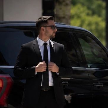 Man in a black suit and sunglasses by a black SUV.