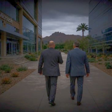 Two men in suits walking between buildings with a mountain backdrop.