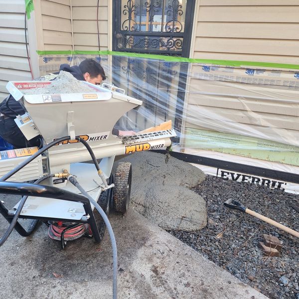 Person using a mud mixer to pour concrete near a house entrance.