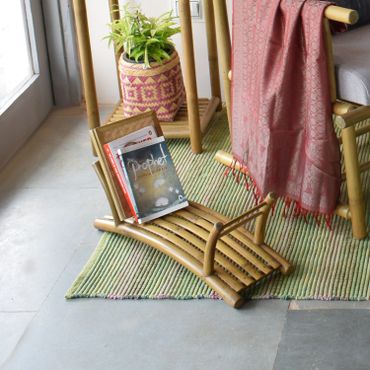 A bamboo footrest with magazines on a striped rug next to a chair and plant.