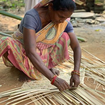 A woman weaving palm leaves into a mat outdoors.