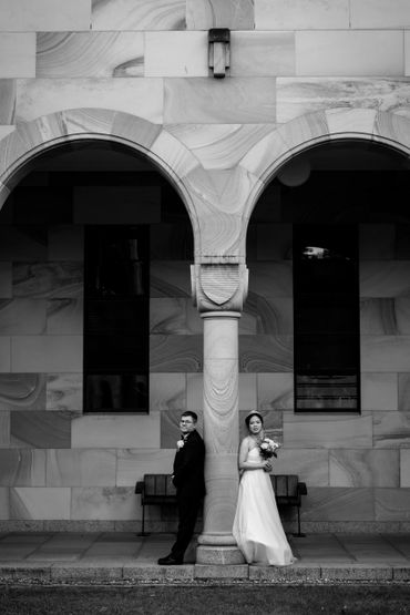 Bride and groom stand back-to-back by a stone pillar under arches.