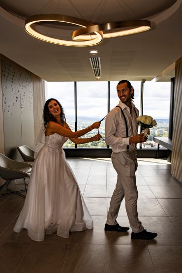 Bride playfully pulls groom's tie, both smiling joyfully indoors.
