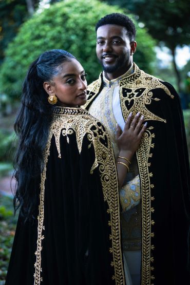 Couple dressed in elegant black and gold embroidered cloaks in a lush outdoor setting.