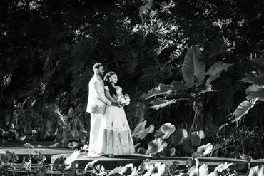 Black and white photo of a couple in wedding attire standing by a pond surrounded by lush foliage.