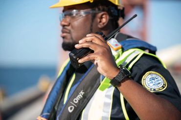 Security guard in helmet and vest using a walkie-talkie at a construction site.
