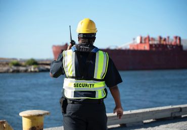 Security guard in high-visibility vest and helmet talks on radio near waterfront.