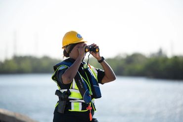 Worker in safety gear observing through binoculars near water.