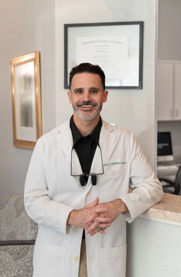 Smiling male doctor in white coat standing in office with diploma on wall.