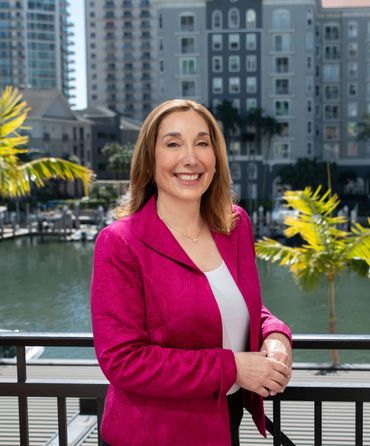 Smiling woman in a magenta jacket standing near waterfront with buildings and palm trees.