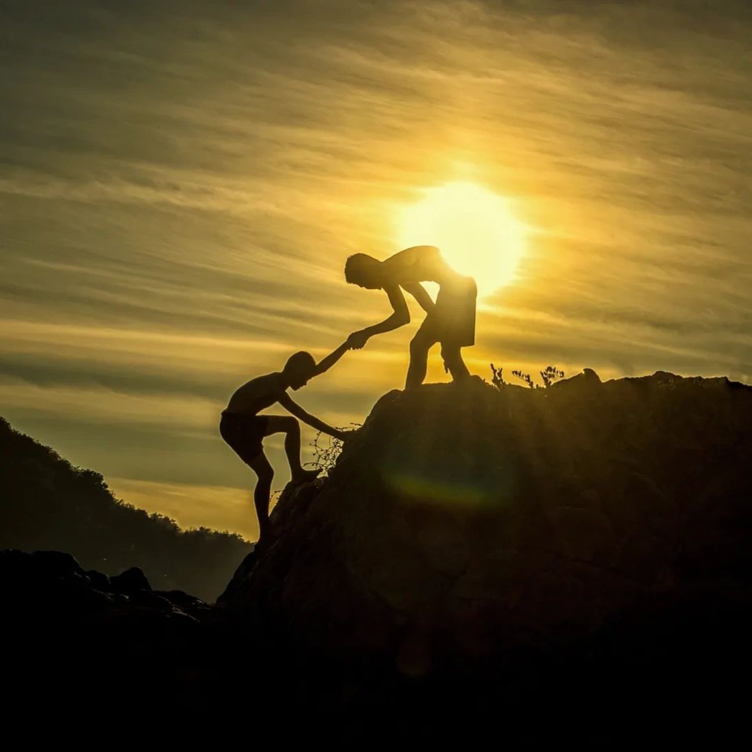 Silhouetted figures at sunset, one helping the other climb a rocky hill.