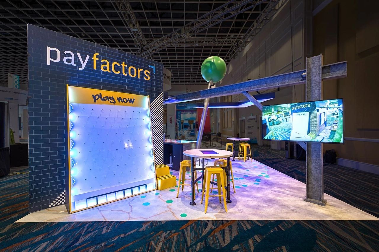 Interactive trade show booth featuring "payfactors" branding, illuminated play area, engaging design elements, and digital display for visitor interaction.