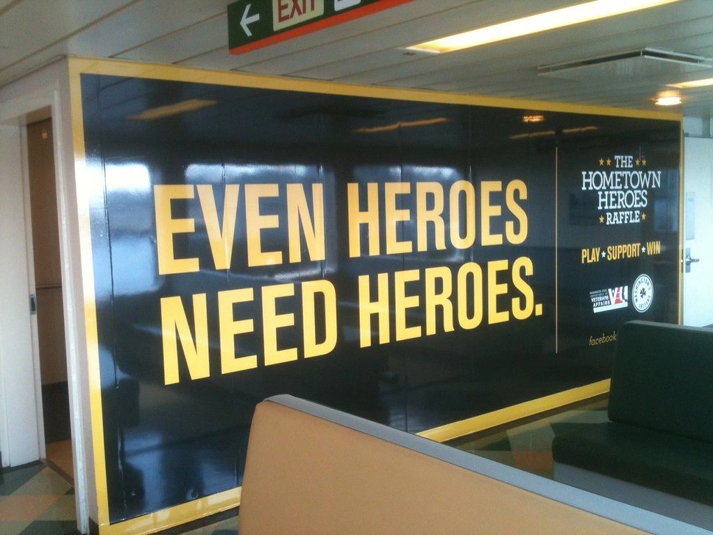 Even heroes need heroes advertisement on ferry wall, promoting Hometown Heroes Raffle with bold yellow text on black background, enhancing commuter experience.